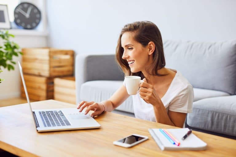 young woman drinking coffee and using laptop while sitting behind table at home