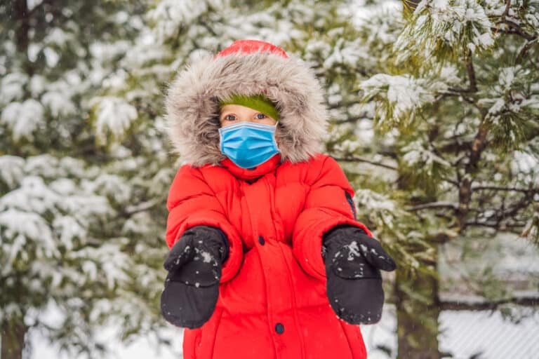 happy boy plays with snow wearing a medical mask during covid 19 coronavirus. cute kid throwing snow in a winter park. happy winter holidays. winter fashion